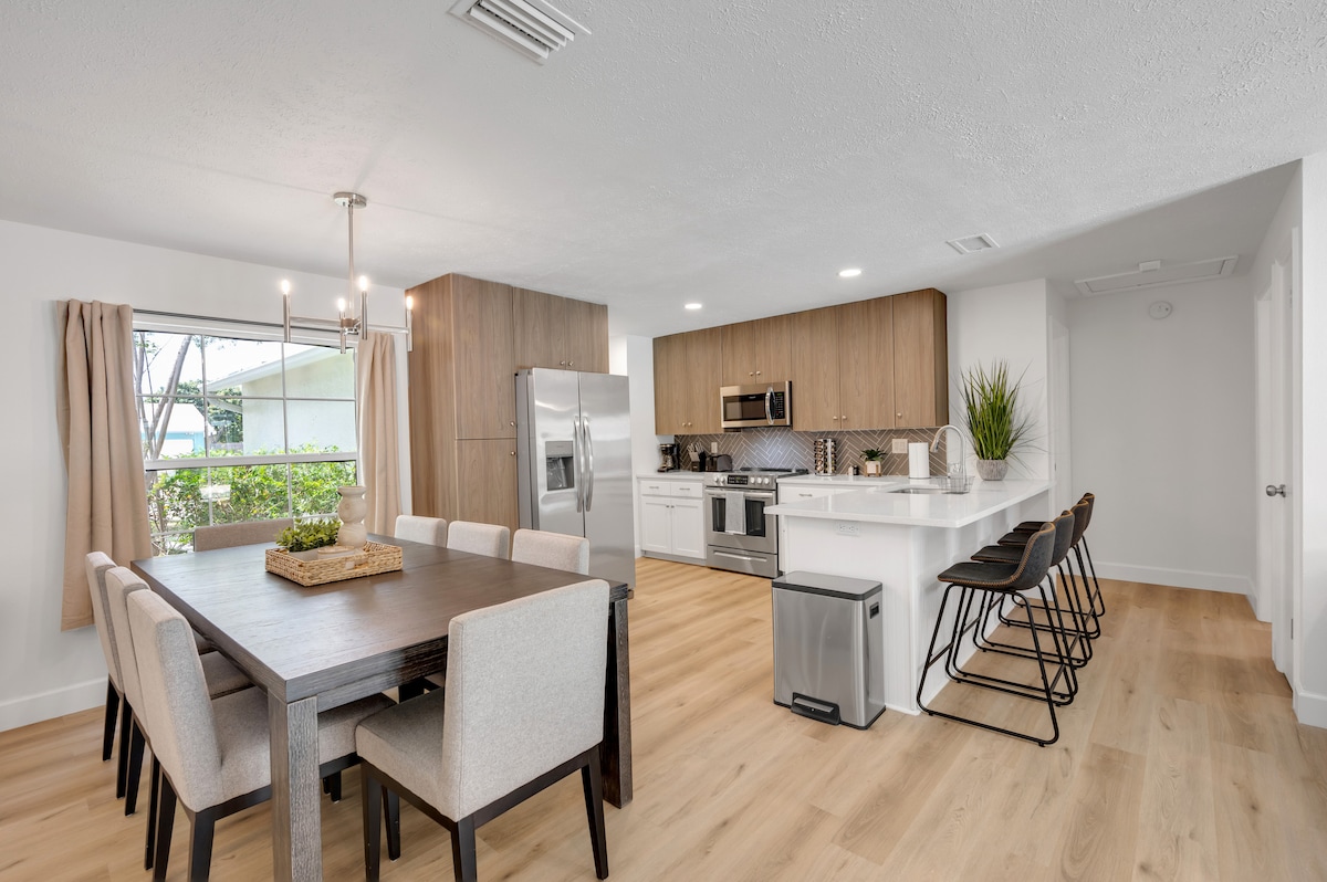 A modern dining area features a wooden table surrounded by upholstered chairs. An open kitchen displays wood cabinetry, stainless steel appliances, and quartz countertops. Large windows allow natural light to illuminate the space, enhancing the inviting atmosphere.