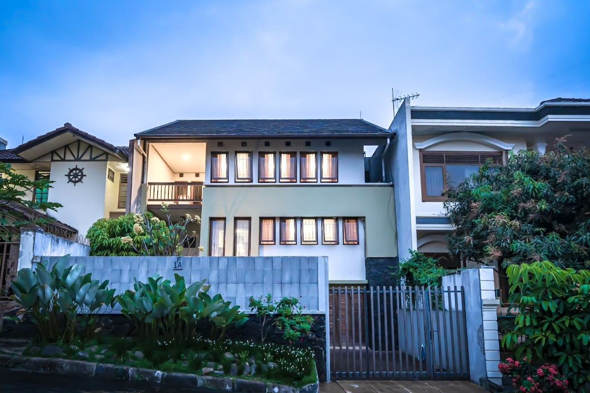 A two-story house with a stone façade features multiple large windows adorned with wooden frames. A spacious balcony is visible on the upper floor, while lush greenery and a gated entrance enhance the welcoming appearance of the property.