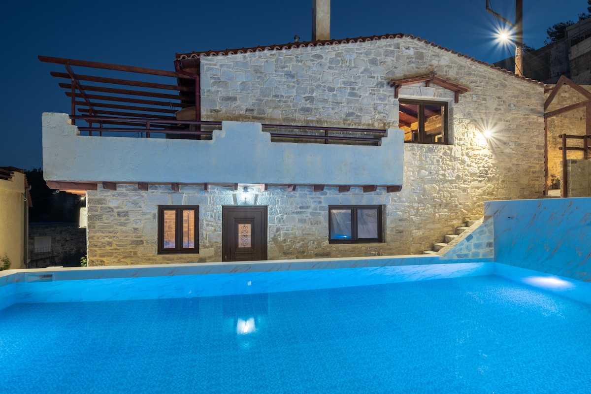 The villa's stone exterior is illuminated at night, showcasing a rustic charm. A spacious pool with clear blue water reflects the surrounding ambiance. Accessible stairs lead to a shaded terrace, enhancing the outdoor living space. Large windows allow for glimpses of the interior.