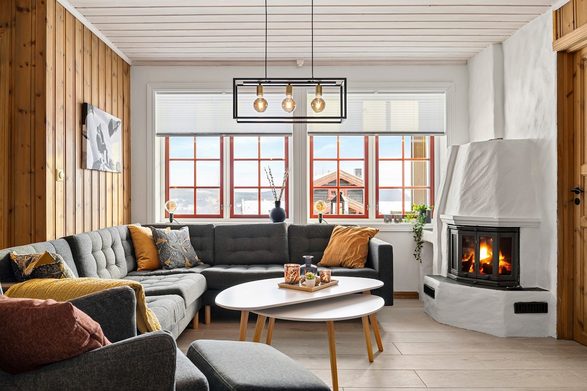 A cozy living area features a sectional sofa arranged around a central coffee table. A fireplace adds warmth, and large windows provide natural light and scenic views. The wooden walls enhance the rustic charm, while decorative pillows and accents contribute to a homely feel.