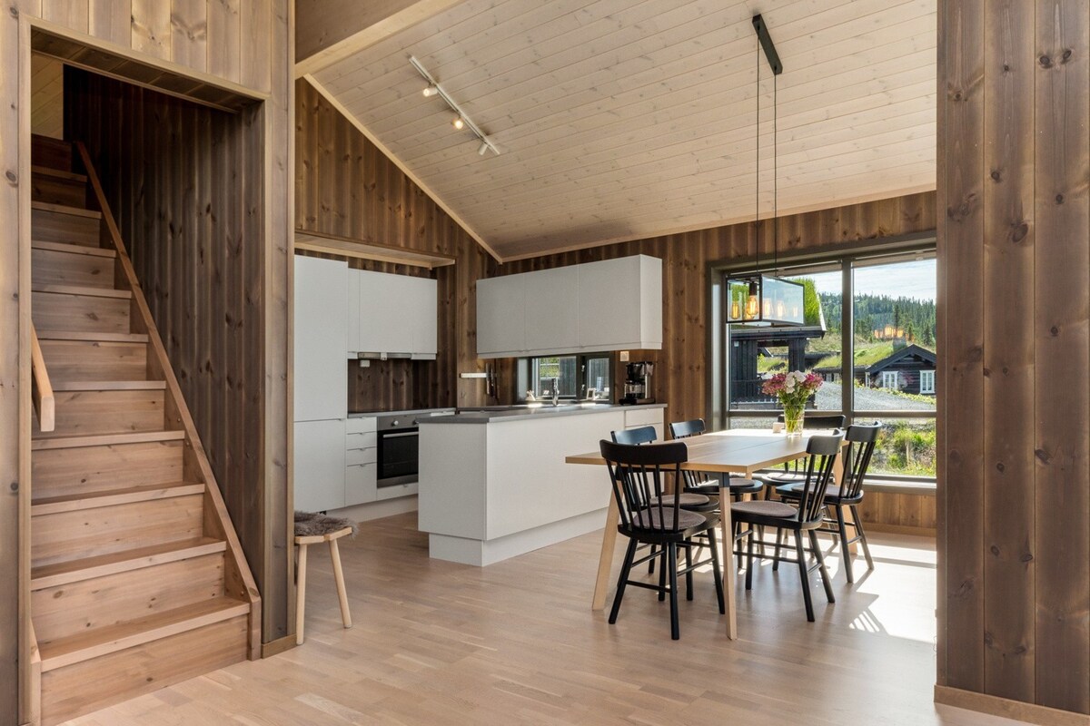 An open-plan kitchen and dining area features light wood walls and flooring. The modern kitchen includes sleek white cabinets and a central island. A dining table with black chairs accommodates several guests, while large windows provide ample natural light and views of the surrounding landscape.