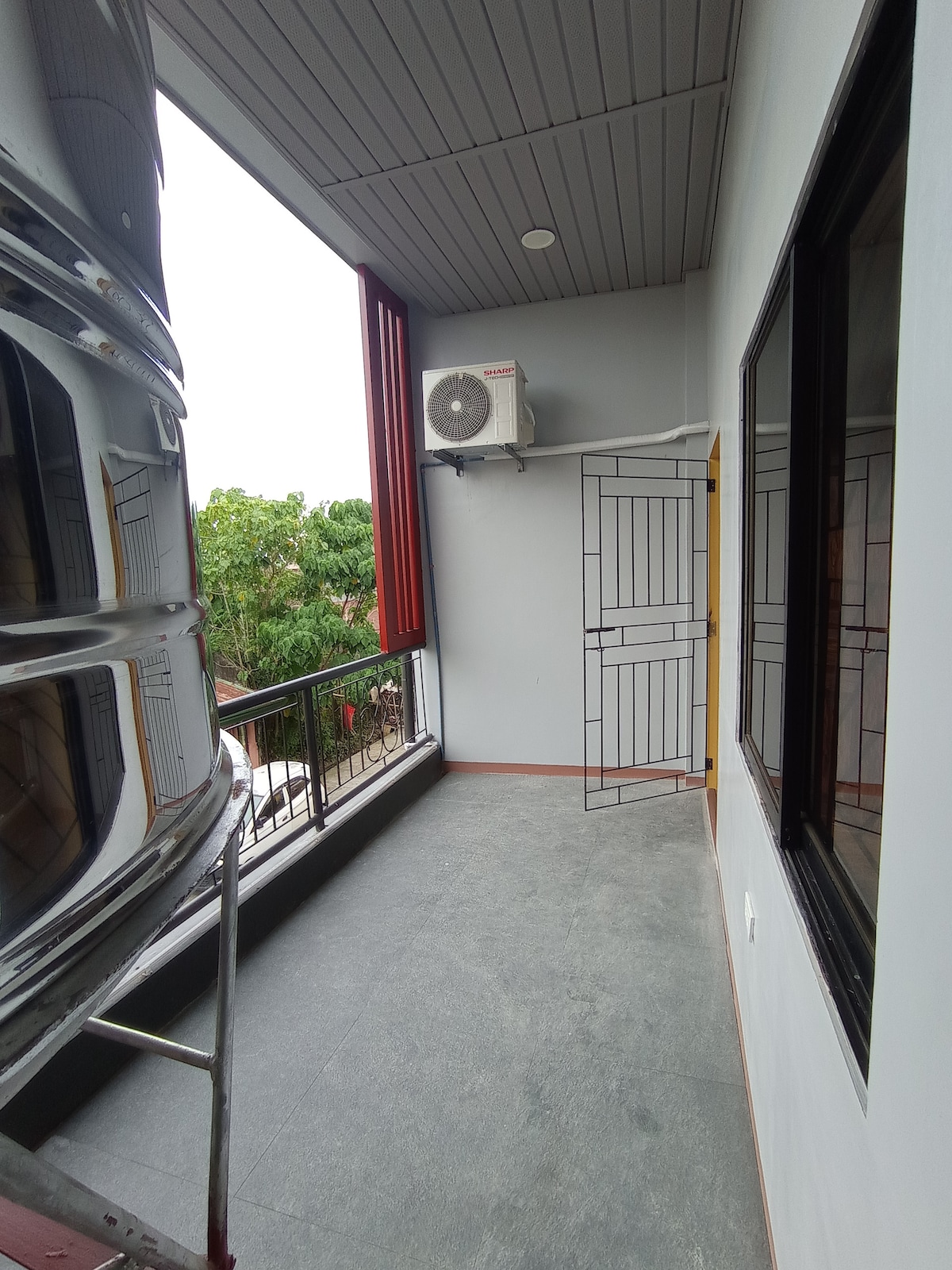 A balcony area features a tiled floor and is bordered by large windows. A wall-mounted air conditioning unit is visible, alongside an iron gate leading to the exterior. Lush greenery can be seen through the openings, providing a refreshing view.
