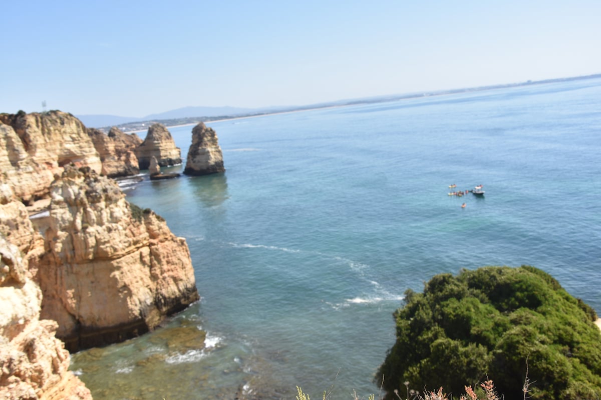 The image captures a coastal scene featuring rugged cliffs rising from a calm blue sea. Small boats can be seen in the distance, and greenery lines the foreground, highlighting the natural beauty of the surrounding landscape.