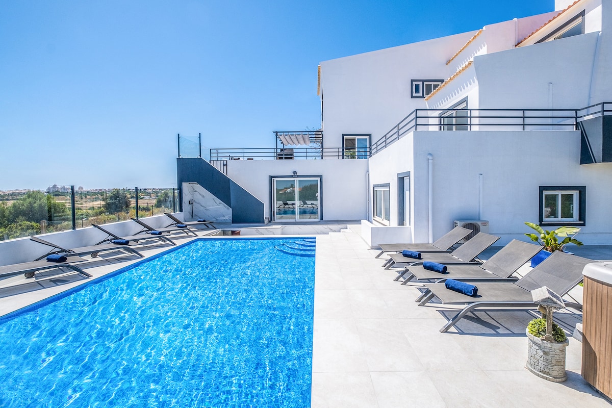 A spacious outdoor area features a vibrant blue pool surrounded by numerous lounge chairs. The villa's stylish white exterior and large sliding glass doors provide a modern backdrop. Views of the surrounding countryside are visible in the distance under a clear blue sky.