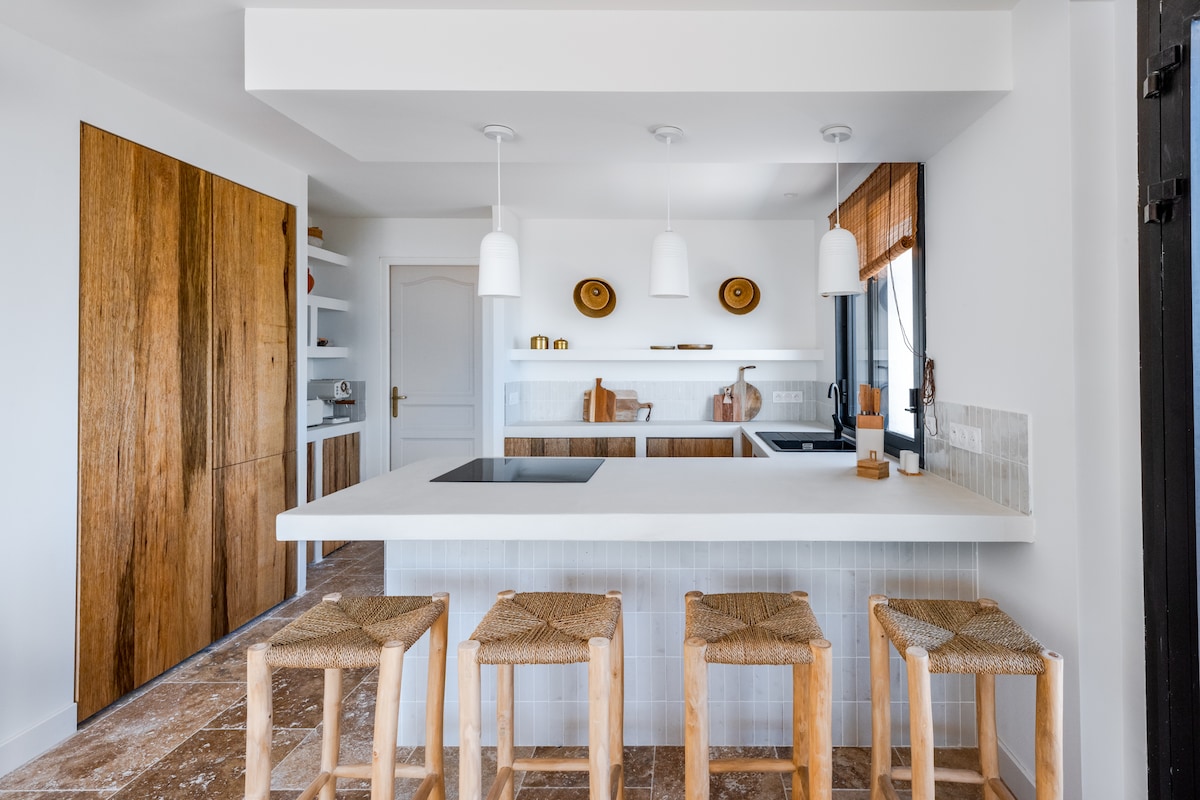 A modern kitchen features an island with a built-in cooktop, surrounded by four wooden stools with woven seats. Light-colored cabinetry contrasts with rich wooden accents, while natural light streams through a nearby window, illuminating the clean lines and minimalist design.