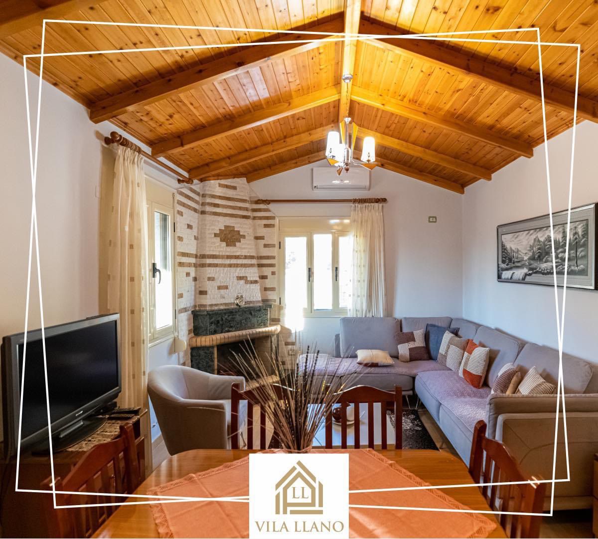 The living area features a high wooden ceiling and is furnished with a sectional sofa and wooden dining table. A television is positioned near the corner, and natural light enters through several windows, creating a bright and inviting space.