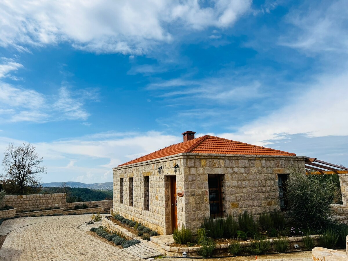 A charming stone house featuring a red-tiled roof is set against a clear blue sky. Surrounding the house, a well-maintained pathway leads to the entrance, with lush green plants and a distant view of the landscape enhancing the serene environment.