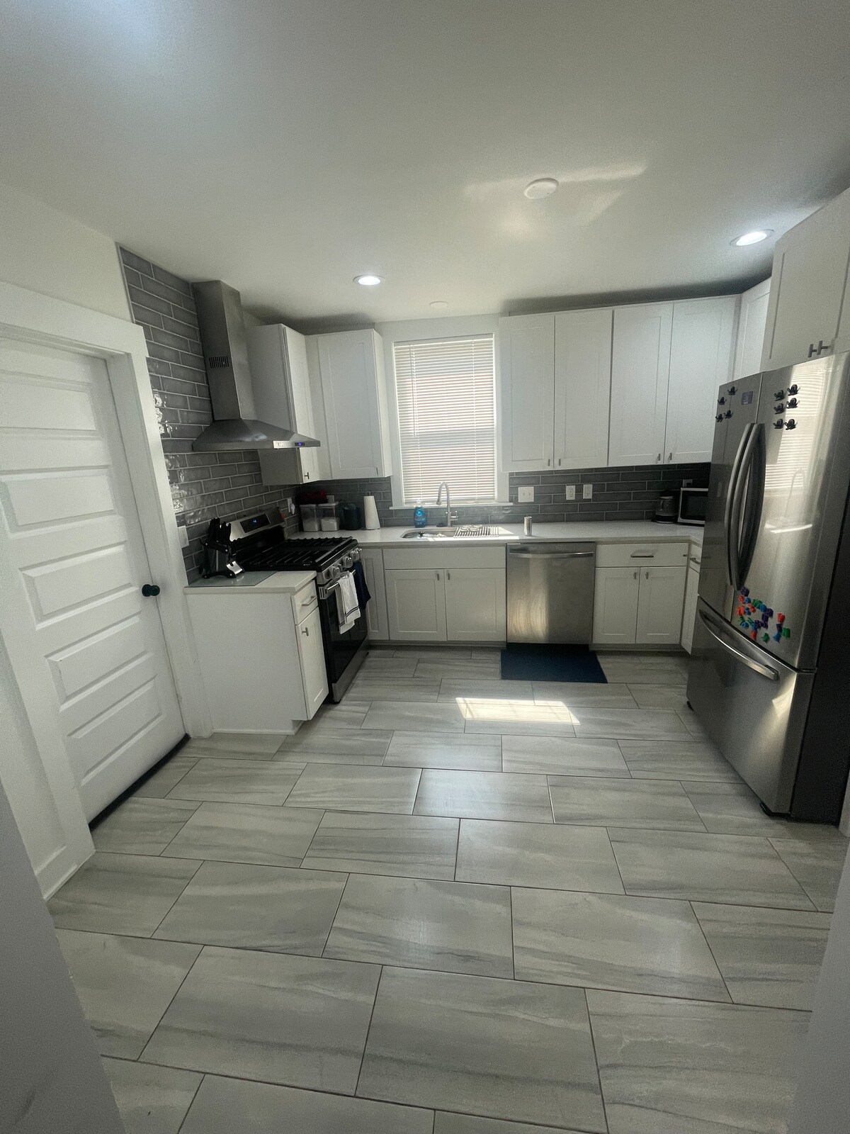 The kitchen features modern white cabinetry and stainless steel appliances, including a refrigerator and stove. A window allows natural light to illuminate the space, which is accented by grey tiled backsplash and a neutral floor tile. The layout is functional and inviting.