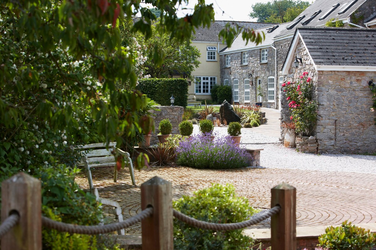 A landscaped courtyard is framed by greenery and flowering plants. Stone buildings with slate roofs form a charming backdrop. A brick path winds through the area, leading to various seating options. Soft hues of lavender and bushes enhance the serene outdoor space.
