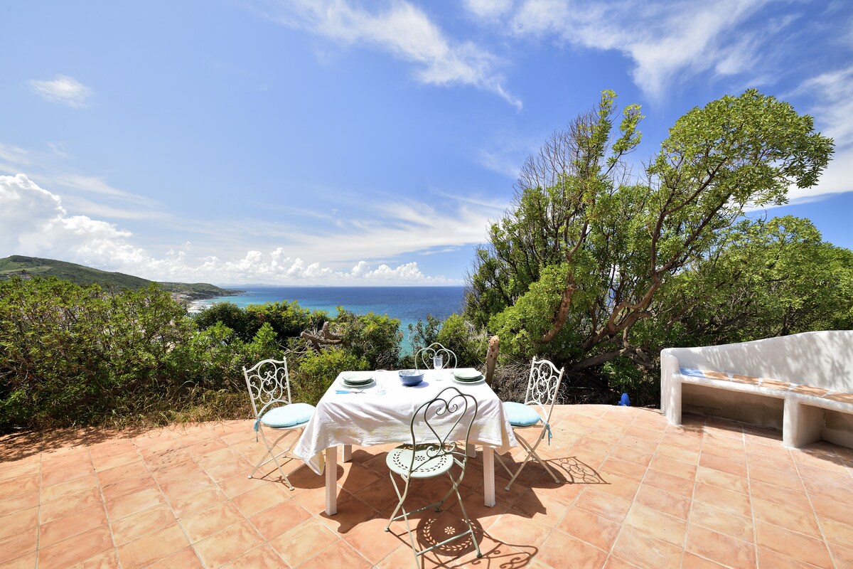An inviting outdoor dining area is set on a tiled terrace, featuring a white table adorned with dishes and utensils. Surrounding greenery frames the scene, while the expansive view of the sea and sky enhances the tranquil ambiance.