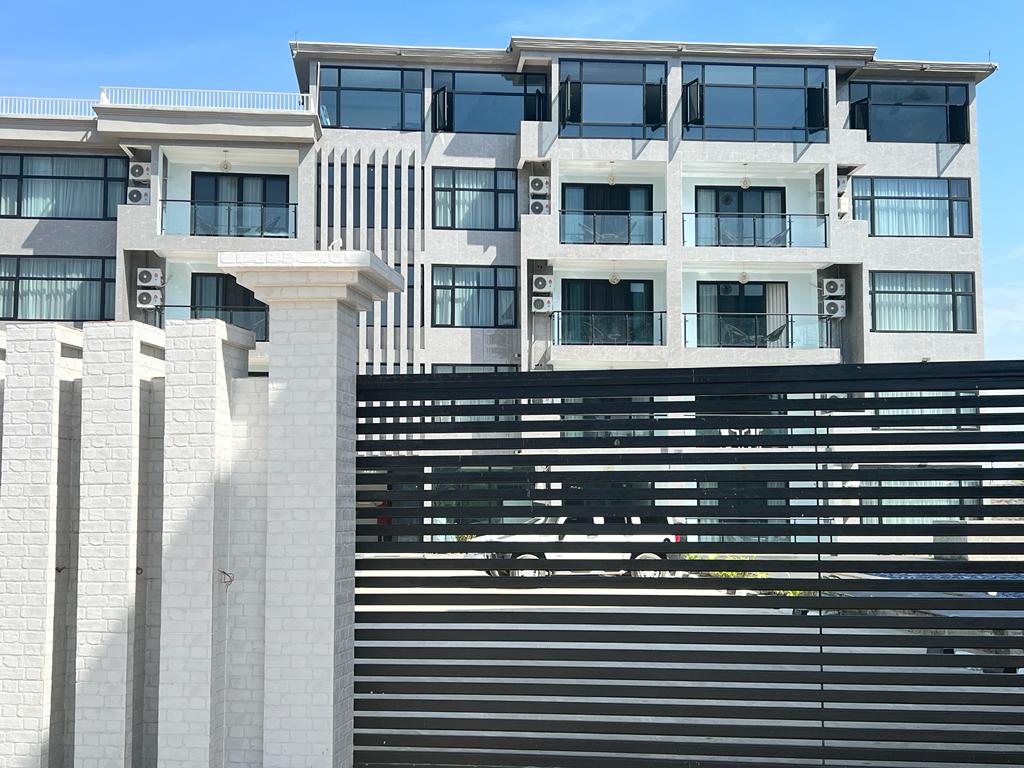 A modern three-story building is presented, featuring large glass windows and multiple air conditioning units. The structure is framed by a contemporary horizontal slat fence, providing privacy while complementing the sleek exterior design.