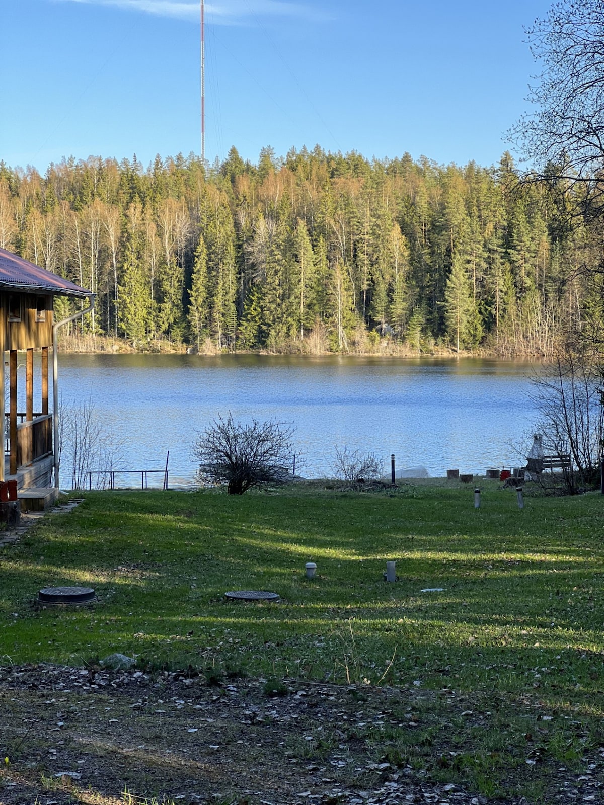 A serene view of the lake is captured, with calm water reflecting the surrounding forest. Lush greenery covers the grassy area in the foreground, dotted with small markers. The backdrop features tall trees that create a natural boundary along the water's edge.