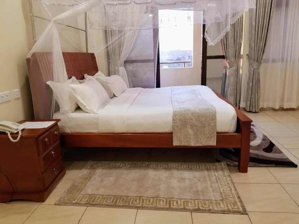 A spacious bed with soft white linens is positioned against a textured headboard. Sheer mosquito netting drapes elegantly around the bed. A wooden bedside table sits nearby, while light-filtering curtains frame the window, creating a serene and inviting space.