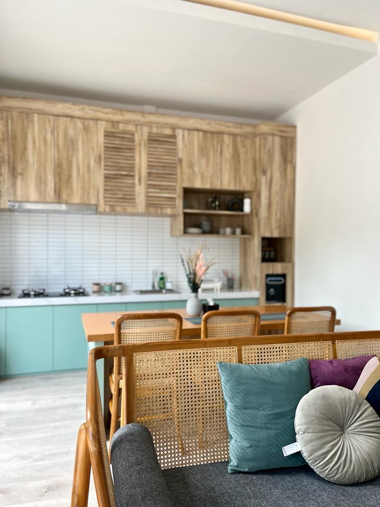 A modern kitchen area is visible, featuring wood cabinetry with slatted details and a light-colored tile backsplash. A dining table with space for four sits prominently, while a cozy couch with a mix of cushions in various colors adds comfort to the living space.