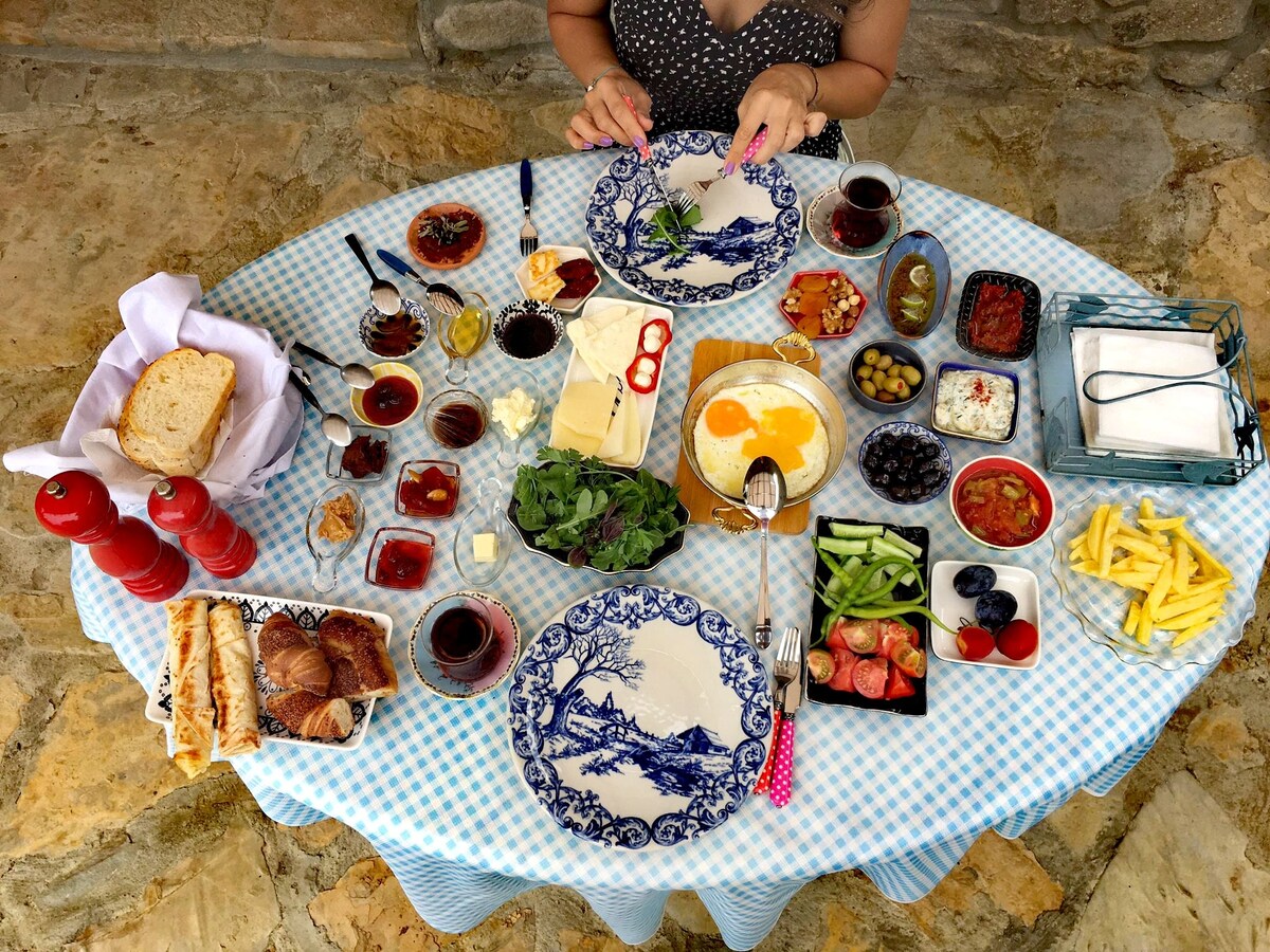 A generously set dining table features a variety of breakfast items, including olives, cheeses, assorted jams, fresh vegetables, and breads. Delicate plates with floral designs are placed at each setting. The table is covered with a light blue checkered tablecloth, creating a welcoming atmosphere.