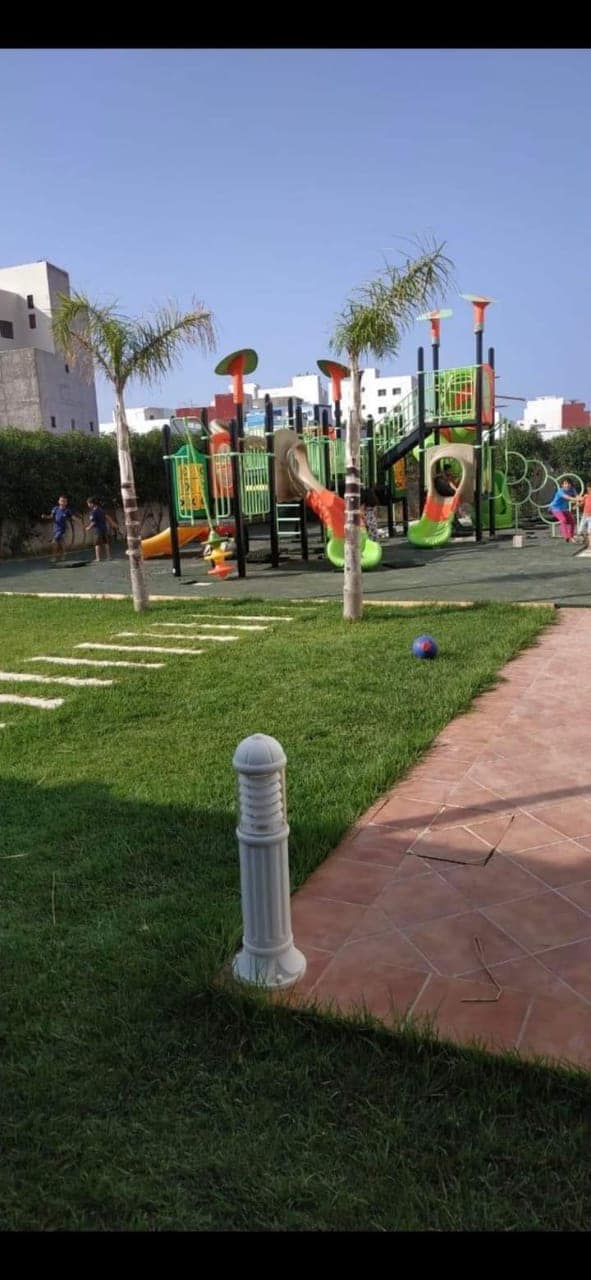 A vibrant playground features a variety of climbing structures and slides surrounded by lush green grass. Colorful play equipment stands adjacent to palm trees, while children can be seen enjoying the space. A pathway made of tiles leads towards the playground area.