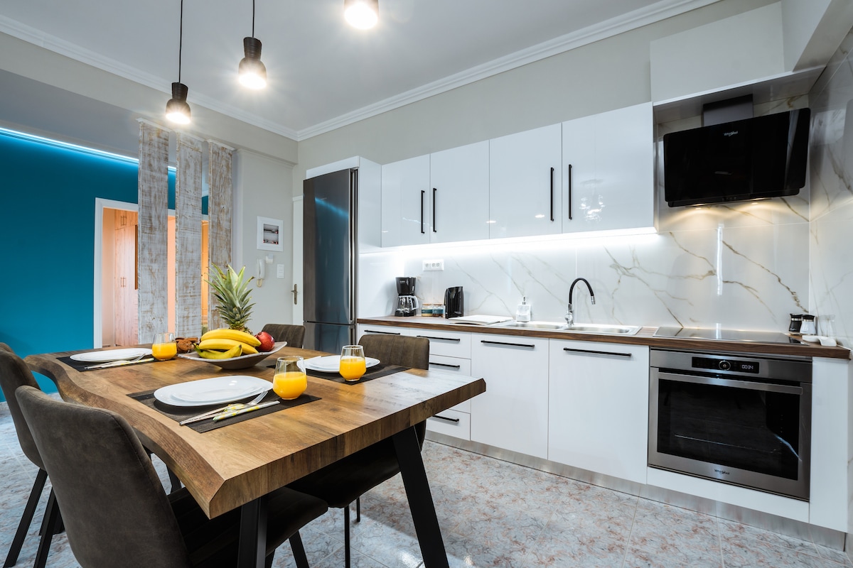 A modern kitchen features sleek, white cabinetry and a marble backsplash. A wooden dining table is set for four, with tablescape elements like plates and drinks. Stainless steel appliances include a refrigerator and an oven, complementing the overall clean and functional design.