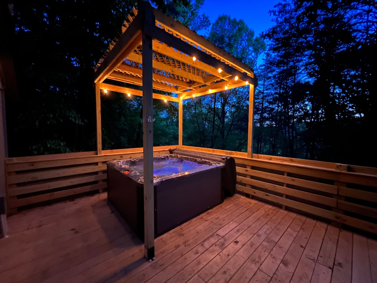 A private hot tub is situated on a spacious wooden deck, illuminated by soft lighting. The surrounding trees create a serene atmosphere, and the structure above offers shade and protection. The deck enhances the outdoor experience, providing a tranquil space for relaxation.