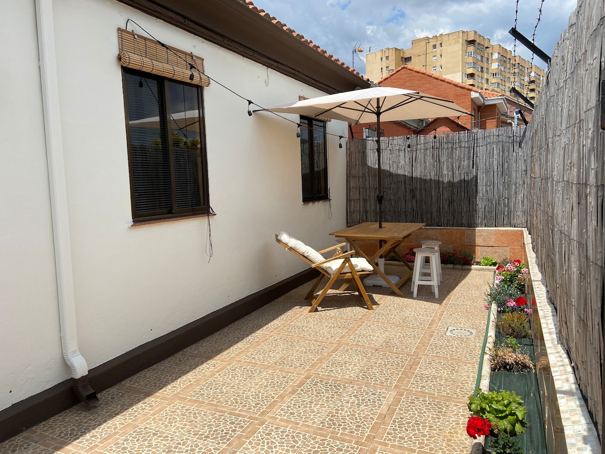 A private outdoor area features a wooden table and chairs, complemented by a large umbrella for shade. The patio is paved with a patterned tile and bordered by low fencing, with flower beds visible along one edge. Natural light fills the space.