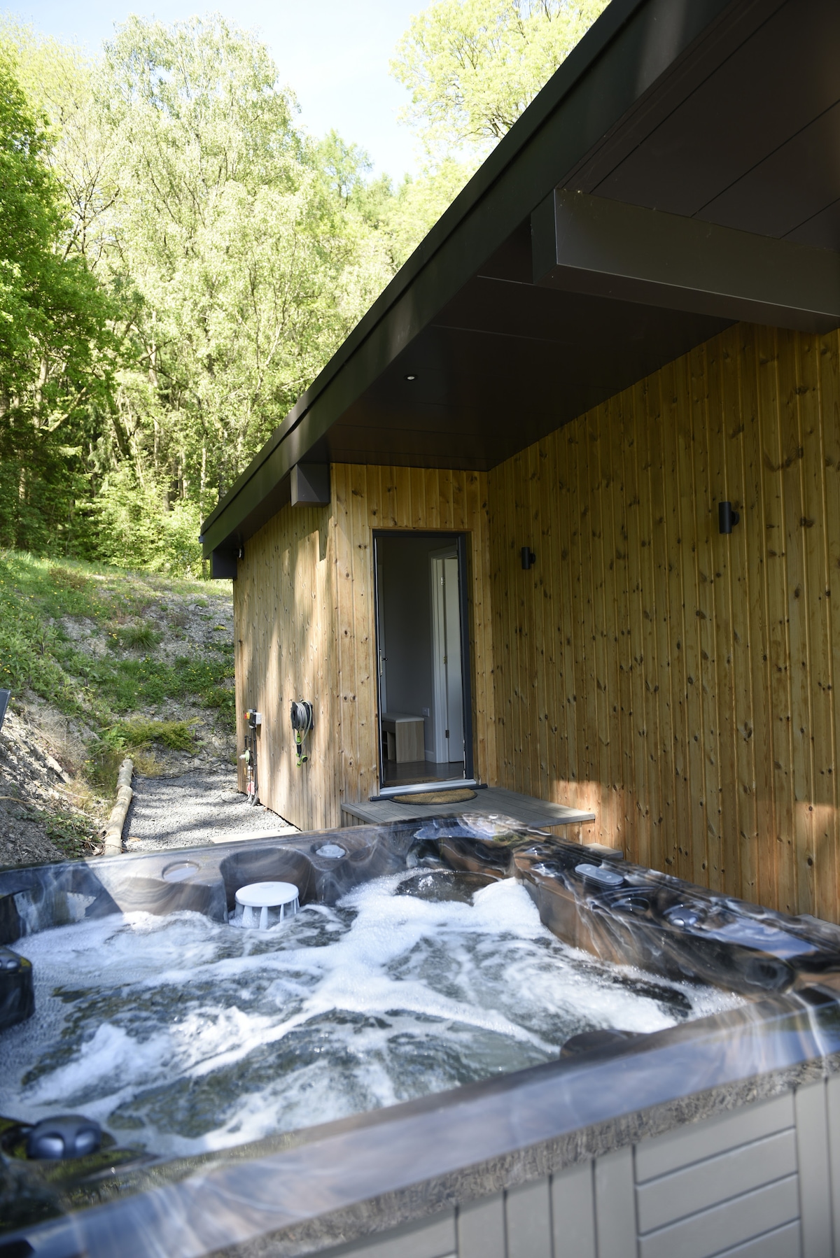 A marquis hot tub is situated on the enclosed veranda, surrounded by natural greenery. A modern wooden structure is visible in the background, featuring large doors that open toward the outdoor space. The area is serene and private, offering a tranquil retreat.