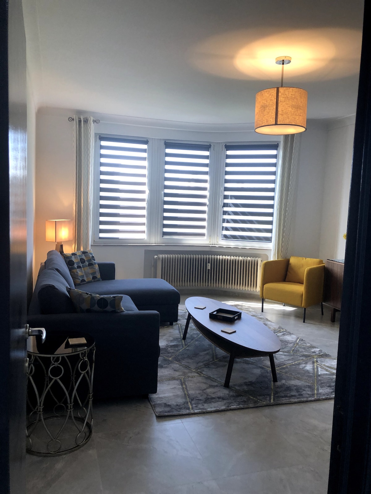 A cozy living area features a blue sofa and a yellow accent chair, positioned around a low coffee table. Large windows with striped shades allow natural light to fill the space. A patterned rug lies under the coffee table, complementing the contemporary design.