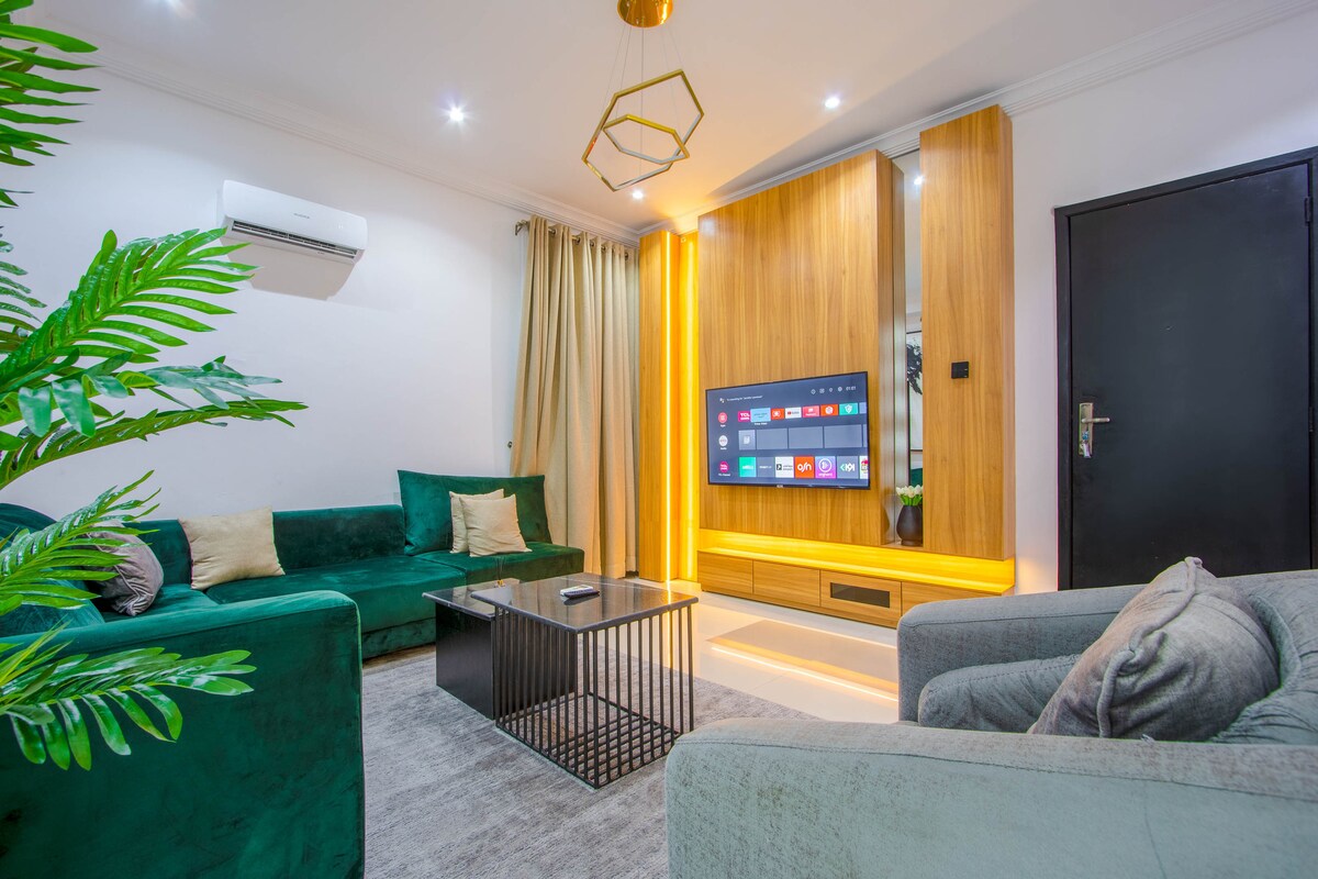 A modern living area features plush green sofas and a cozy gray couch. A sleek coffee table sits in the center, while a wall-mounted television is surrounded by warm wooden accents and ambient lighting. Large windows are dressed in soft curtains.