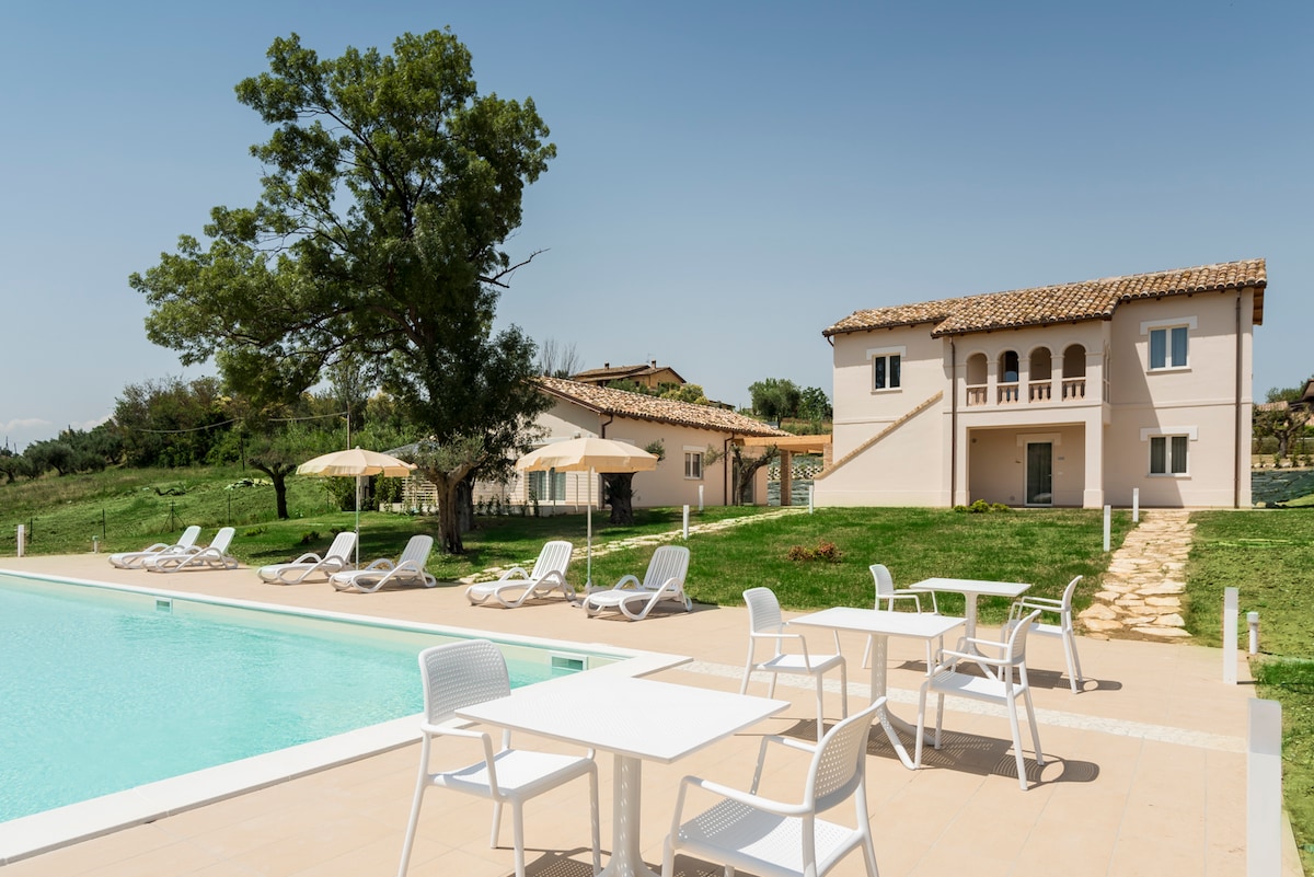 A tranquil outdoor area features a large swimming pool surrounded by sun loungers and tables. The inviting structure shows a balcony and several large windows, set against a backdrop of lush greenery and a clear blue sky.