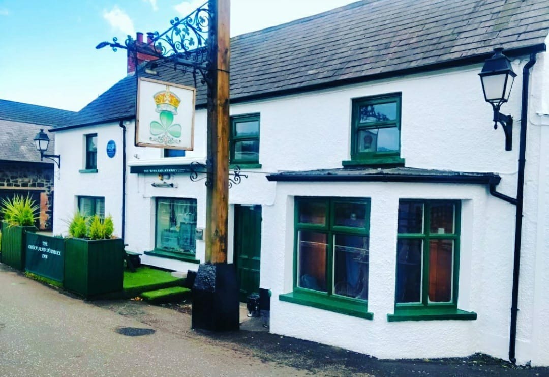 The exterior of a traditional Irish pub is displayed, featuring a white façade with green trim. The signage includes a crown and clover motif, while decorative lanterns frame the entrance. A patch of grass is visible in front, contributing to the welcoming atmosphere.