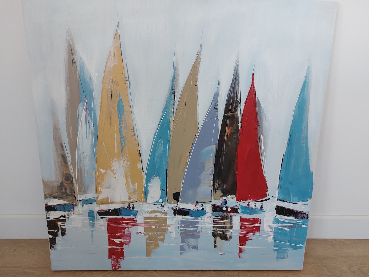 An abstract painting depicting various sailboats with sails in shades of blue, red, yellow, and brown. The boats are reflected in calm waters, while a light background enhances the colors. The artwork is framed simply, adding to the overall aesthetic.