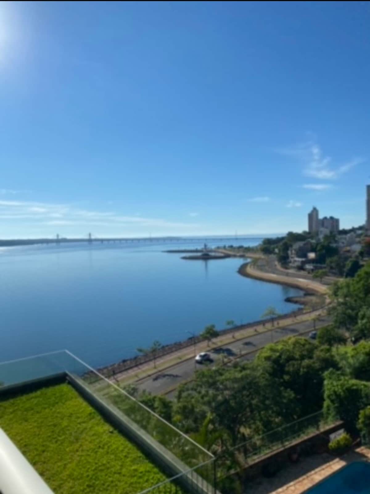 A panoramic view showcases a serene river landscape, with lush greenery lining the shore. The calm water reflects the blue sky, while a distant bridge and city skyline are visible in the background, suggesting a tranquil environment along the waterfront.
