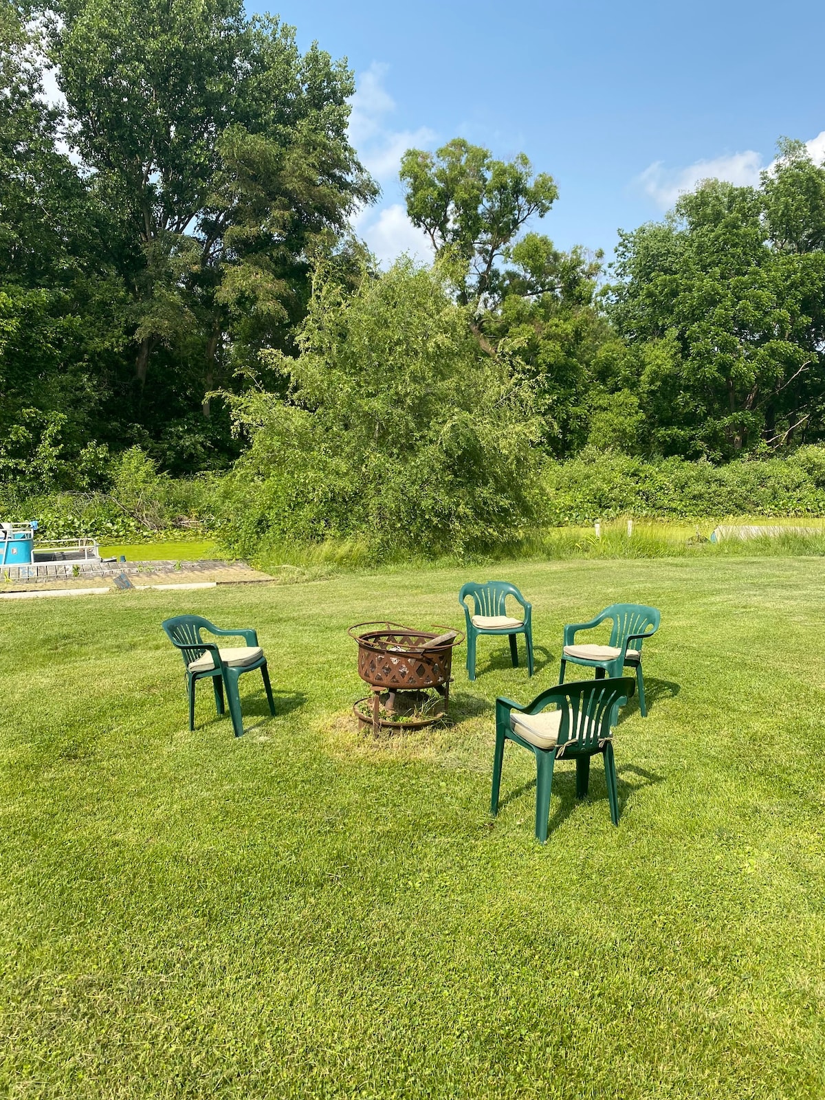 A circular arrangement of green patio chairs surrounds a fire pit on a grassy area. Lush greenery and trees provide a natural backdrop, while warm sunlight brightens the scene. The fire pit is centrally located, inviting companionship and relaxation.