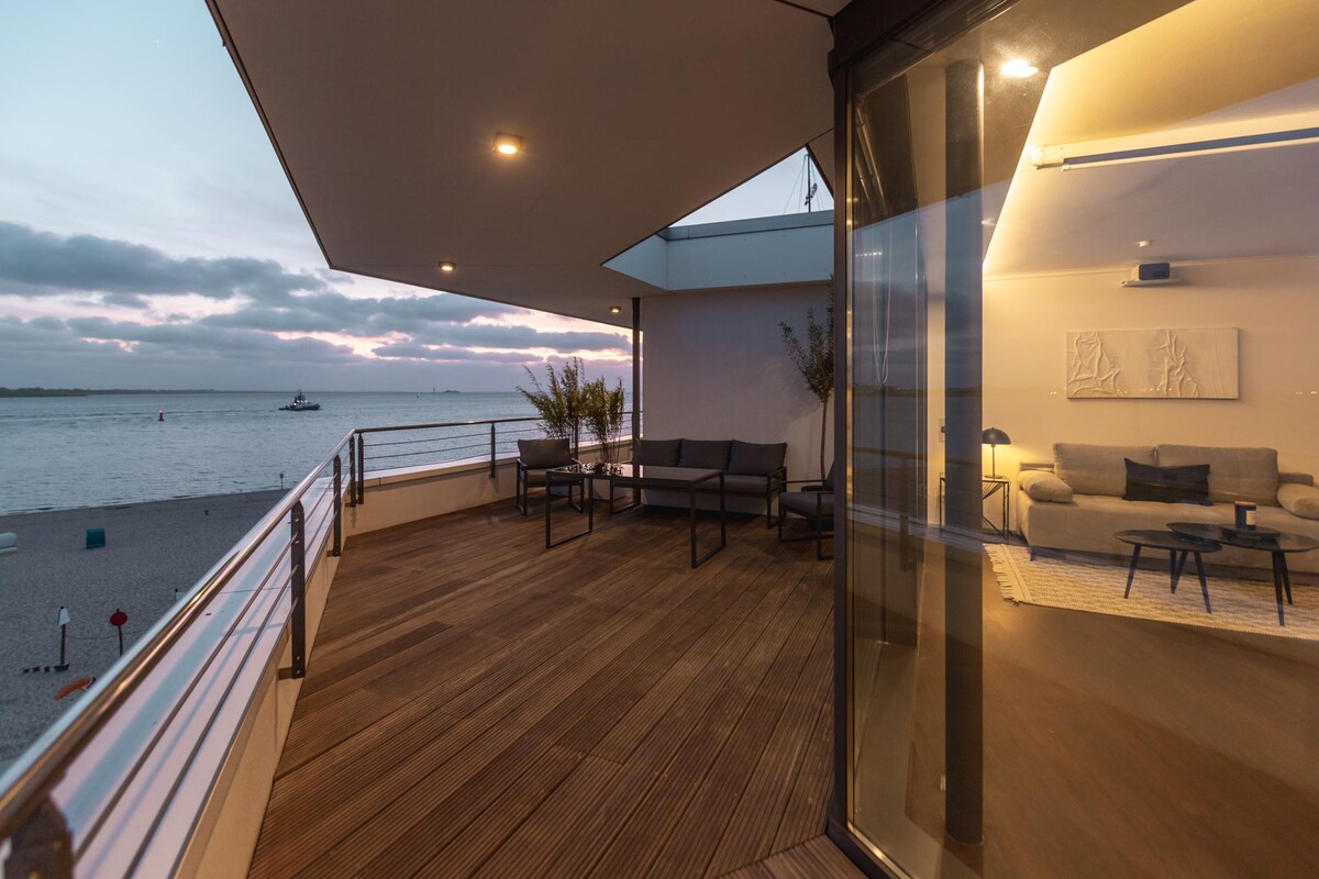 A spacious terrace is shown, framed by large glass doors. Seating arrangements are visible, overlooking the water with muted sunset hues in the background. The interior, softly lit, features a comfortable couch, offering a seamless transition between indoor and outdoor spaces.
