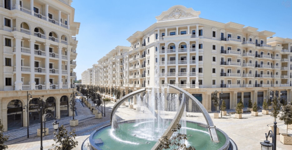 The exterior of a modern residential complex features elegant architecture with multiple balconies and large windows. A central fountain operates, surrounded by carefully arranged landscaping. The streets are lined with trees, contributing to a serene urban environment.