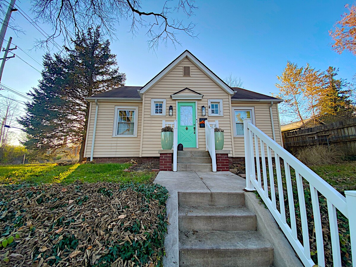 The charming exterior of the home showcases a light-colored facade and a welcoming front porch. Two large planters flank the entrance, which features a mint green door. The steps lead to a well-maintained yard, surrounded by greenery and trees.