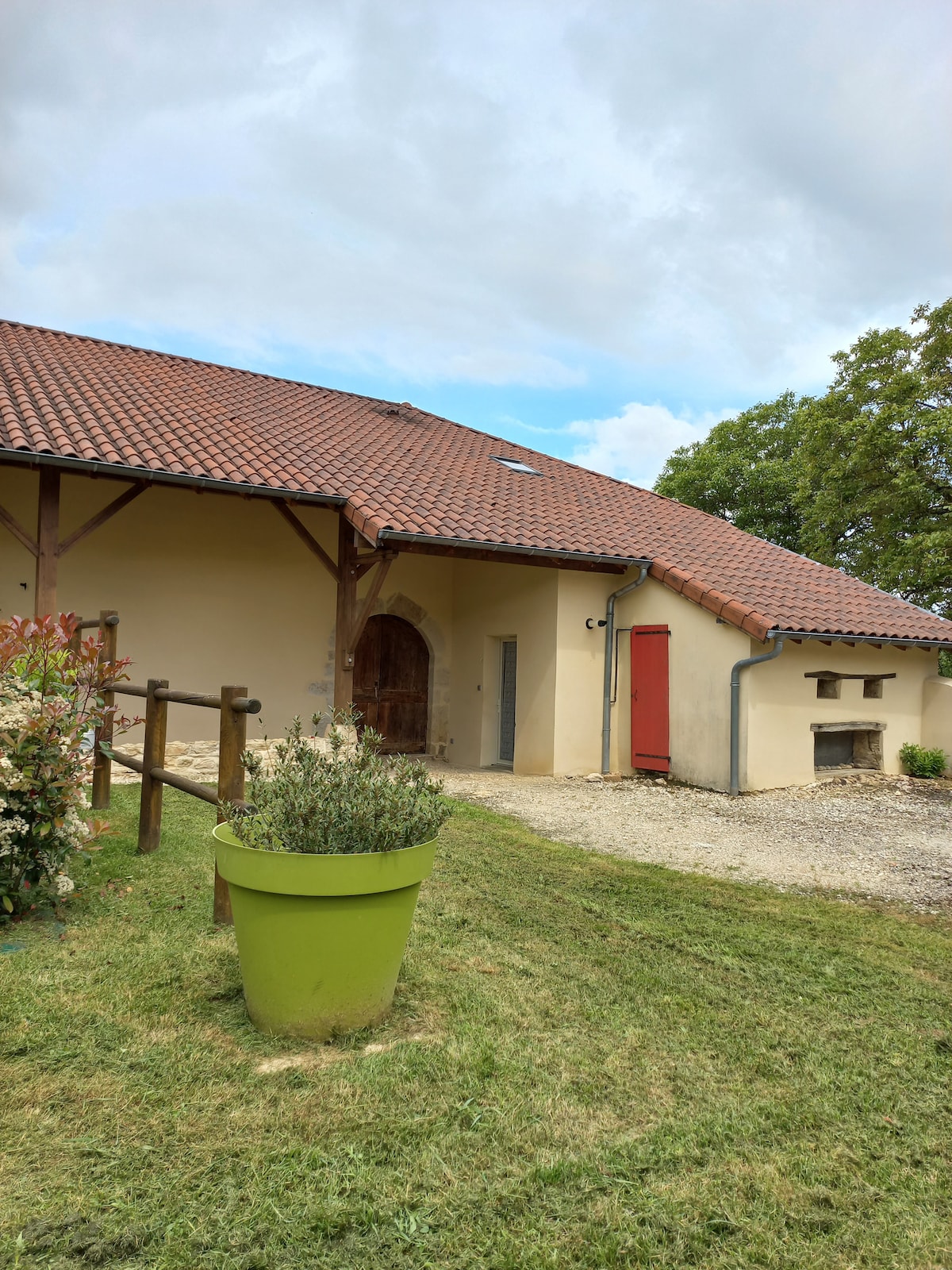 A tranquil exterior view of the building showcases a countryside charm with a red door and tiled roof. Lush greenery surrounds the area, including a large, vibrant green planter filled with plants. A gravel pathway leads to the entrance, emphasizing the peaceful setting.