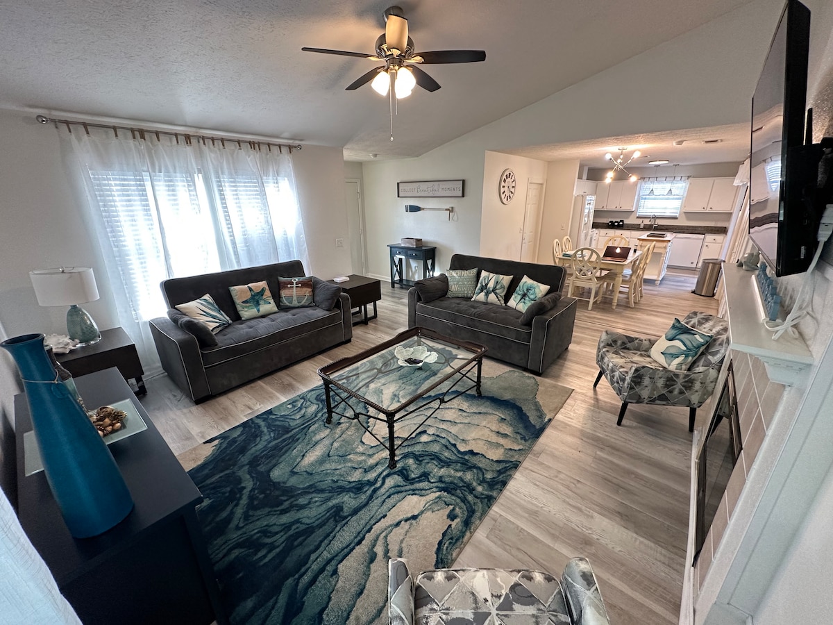 A spacious living room is shown, featuring two sofas, a patterned chair, and a coffee table centered on a textured area rug. Large windows allow natural light to fill the room, complemented by ceiling fans and neutral walls. A dining area is visible in the background, showcasing a white table and chairs.