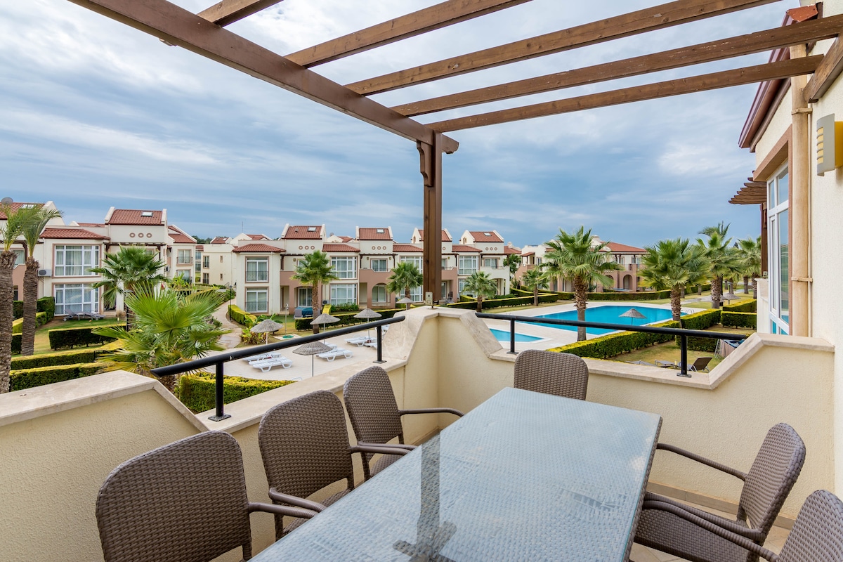 A spacious outdoor terrace is presented, featuring a glass-top table surrounded by six woven chairs. Lush palm trees and landscaped gardens are visible in the background, along with a swimming pool. Overhead, a wooden pergola provides shade, enhancing the outdoor experience.