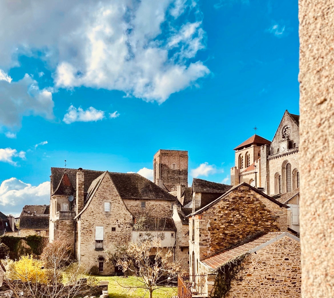 A view of historic stone buildings under a bright blue sky with scattered clouds. The scene includes a mixture of structures, showcasing architectural diversity. A church tower is prominent in the background, while greenery peeks through the spaces between the buildings.