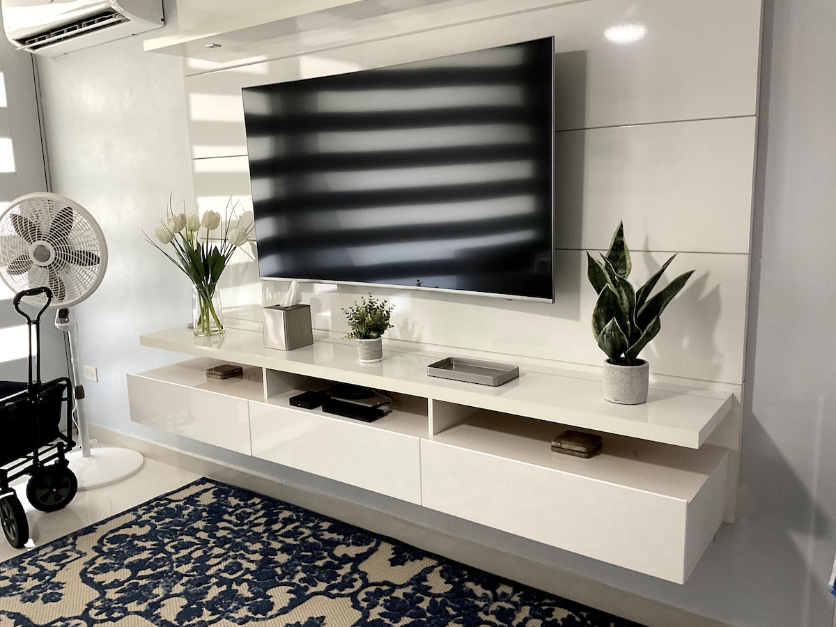 A wall-mounted television is positioned above a sleek, white media console. Potted plants and decorative items are placed on the console, while a patterned area rug lies beneath. Natural light filters through the window, creating a bright atmosphere complemented by the ceiling fan nearby.