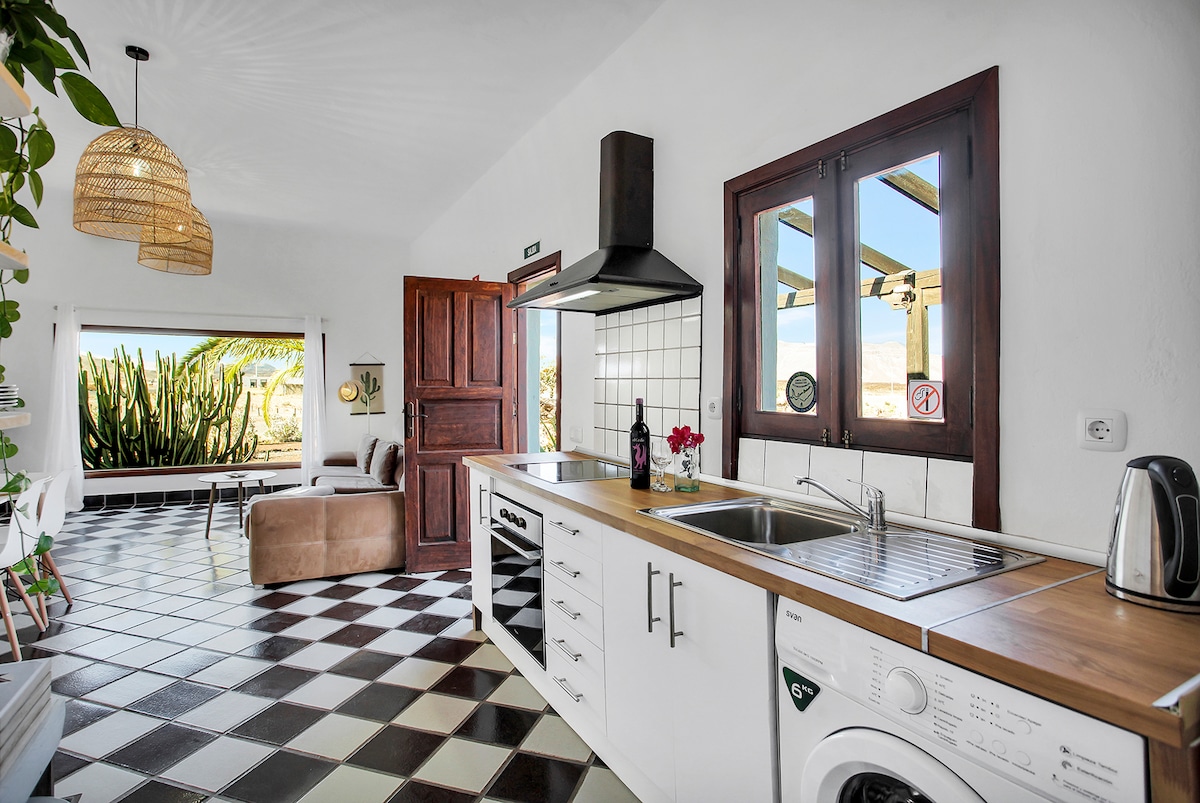A bright kitchen area features modern appliances, including a sink, washing machine, and kettle, set against a backdrop of tiled flooring. A window offers views of the surrounding landscape with cacti, while natural light fills the space through an adjacent entryway.