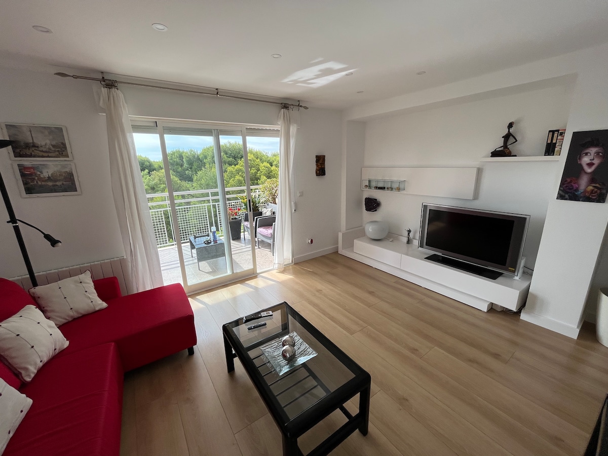 A bright living room features a red sofa and a glass coffee table. Natural light fills the space through large sliding doors that open to a terrace, revealing views of greenery. A minimalist entertainment unit sits against the wall, complemented by decorative accents.