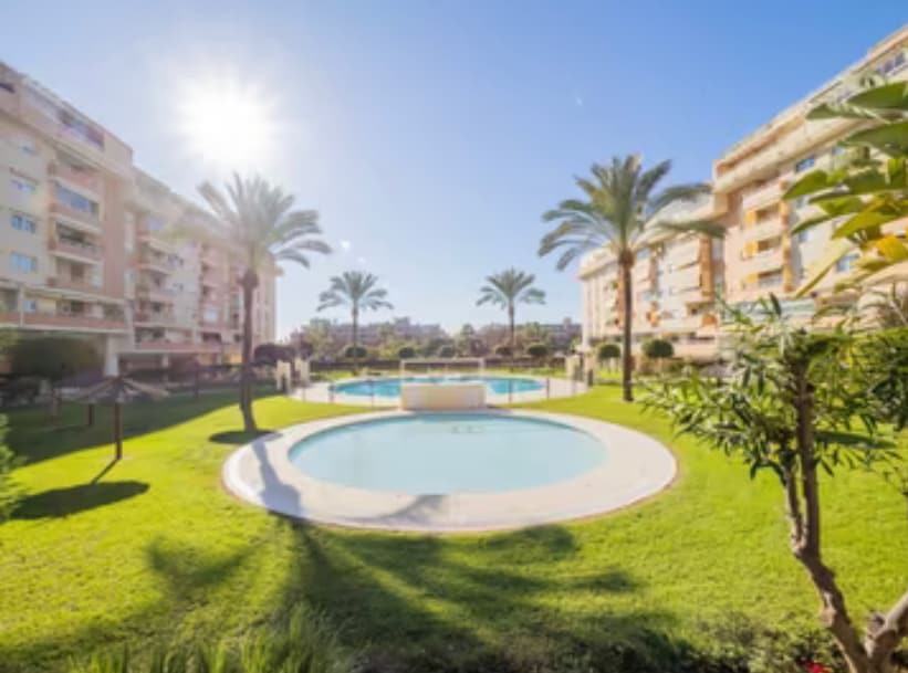 The serene outdoor area features lush green lawns surrounded by palm trees. A circular pool sits at the center, reflecting sunlight. Apartment buildings are visible in the background, providing a sense of community. The space is well-kept and inviting, perfect for relaxation.