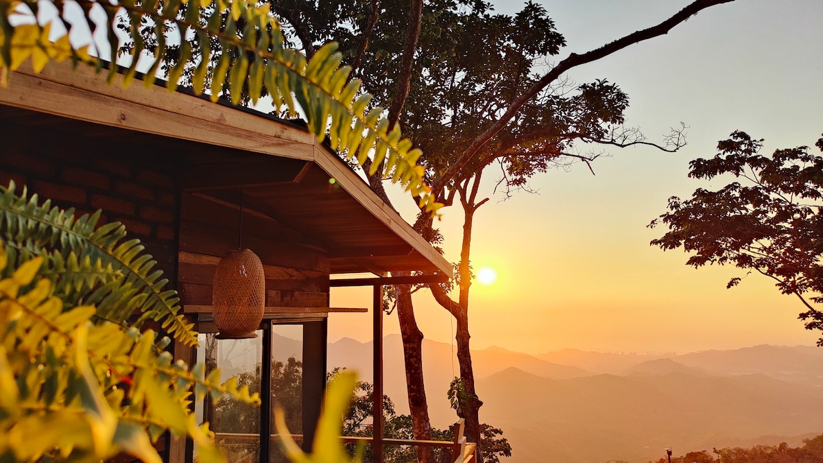 The image captures a cozy bungalow nestled among trees, framed by vibrant ferns. A warm sunset casts light over the surrounding mountains, enhancing the serene landscape. The structure features large windows, inviting in the natural beauty of the environment.