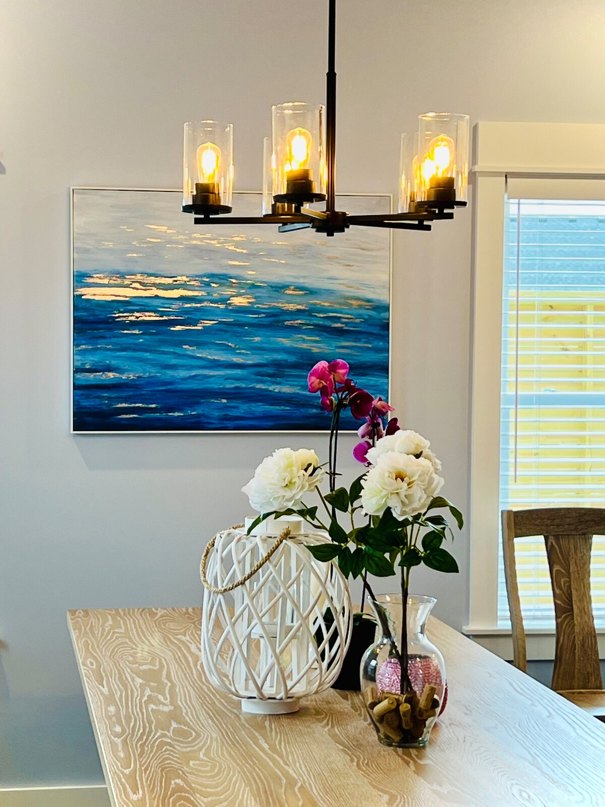 A contemporary dining area is highlighted by a modern chandelier with four lights. A large canvas painting depicting a blue ocean scene adorns the wall, while a vase filled with white flowers and an orchid sits on a wooden dining table.