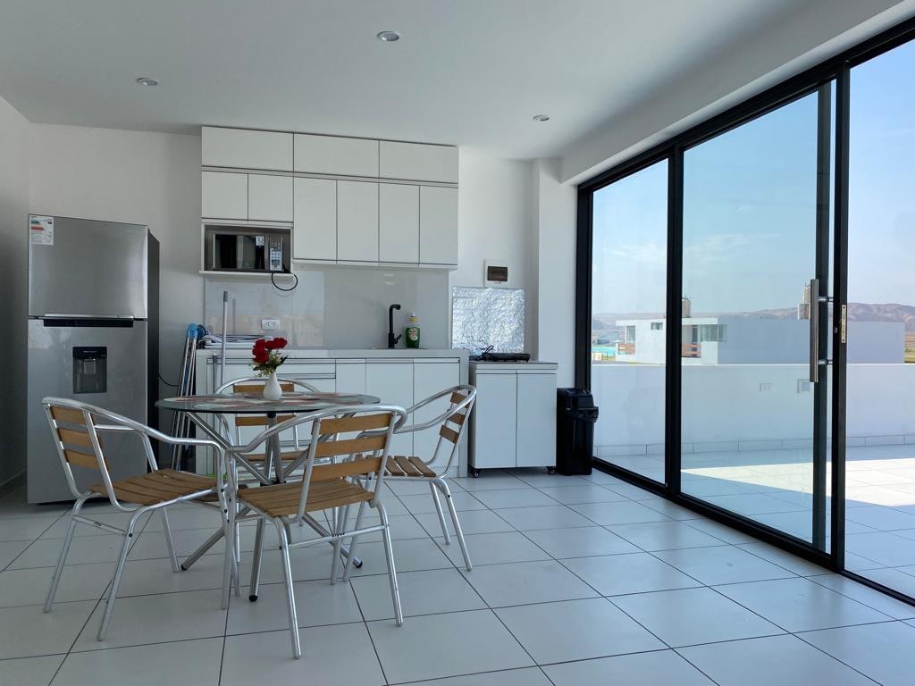 A modern kitchen space features white cabinetry and stainless steel appliances, including a refrigerator and stove. A dining table with four bright wooden chairs sits centrally, illuminated by natural light from a large glass door that opens to the balcony.