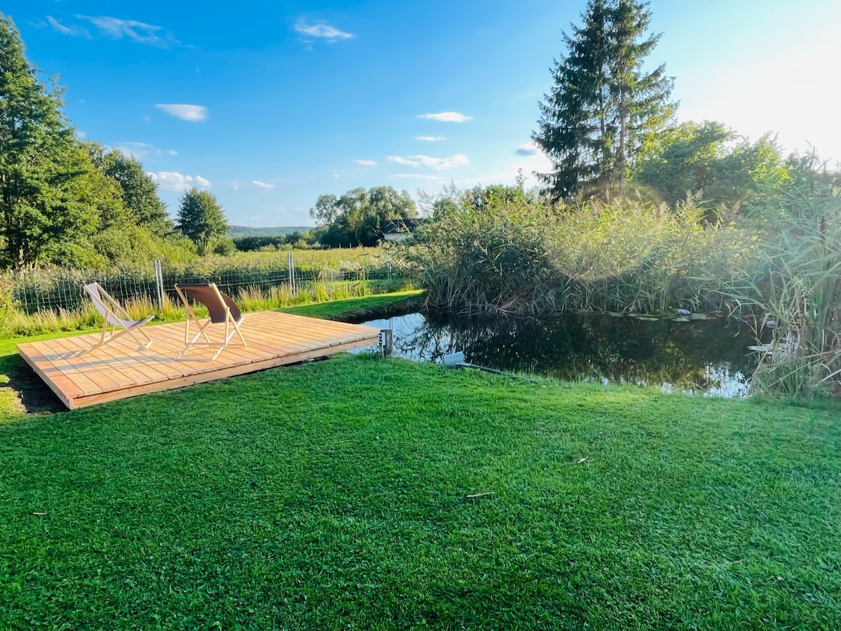 A wooden deck overlooking a small garden pond is surrounded by lush green grass and tall reeds. Two reclining chairs are positioned on the deck, inviting relaxation while enjoying the serene natural landscape under a clear blue sky.