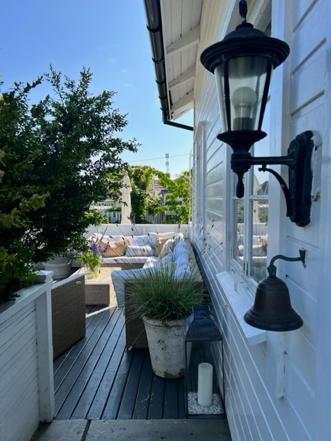 An inviting outdoor seating area is set on a wooden deck, surrounded by greenery. Comfortable seating arranged around a central space fosters relaxation. A decorative lantern is mounted on the exterior wall, enhancing the serene atmosphere of the garden.