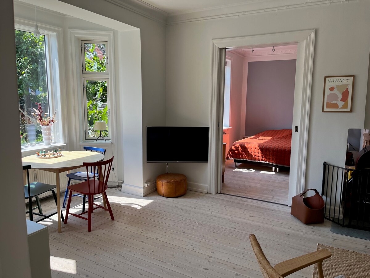 A bright and open living space is displayed, featuring a small dining table with colorful chairs. A flat-screen TV is mounted on the wall, and a view into a cozy bedroom is visible. Natural light fills the room, highlighting the warm wooden flooring.