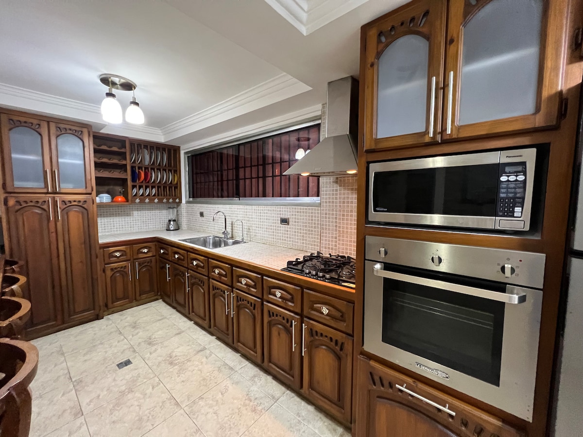 A fully equipped kitchen is displayed, featuring wooden cabinetry and a light tile backsplash. Stainless steel appliances, including a microwave and oven, complement the cooking area. Natural light is provided by a window, while ample counter space supports meal preparation.