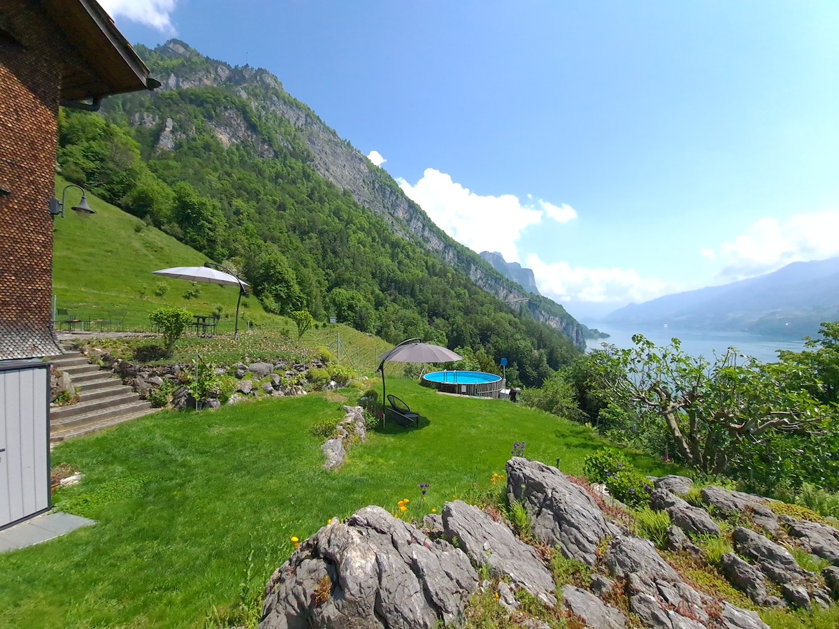 An expansive green lawn is bordered by rocky terrain, leading to a swimming pool surrounded by sun loungers and umbrella shade. Majestic mountains rise in the background, with views of the lake creating a serene natural setting under a clear blue sky.